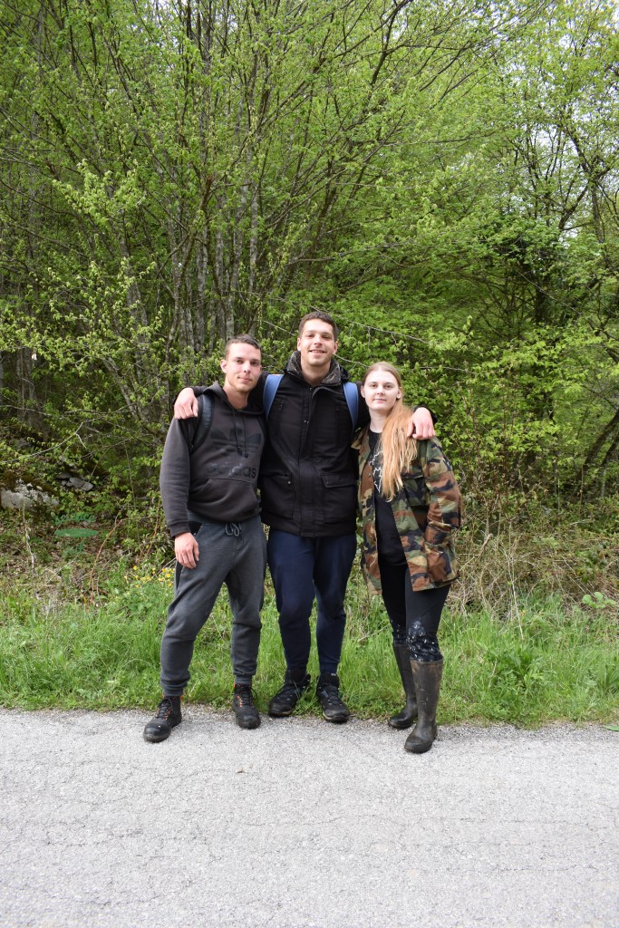 Three people in embrace (two men, one woman) posing, on the road in front of the trees and thicket.